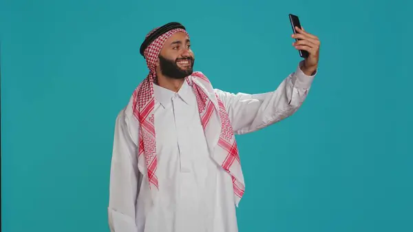 stock image Young adult taking photos with phone, capturing memories and funny faces on smartphone camera over blue background. Middle eastern guy with checkered headscarf takes pictures in traditional attire.