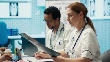 Tıbbi uzman röntgen sonuçlarını inceleyerek, sağlık personeli ve tedavi seçenekleriyle yaptığı toplantıda hastalıkların önlenmesini tartışıyor. Radyoloji doktorları teşhis taramalarını inceliyor. Kamera A.