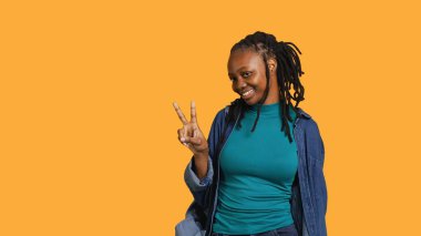 Neşeli Afro-Amerikan genç bayan gülümsüyor, zafer işareti yapıyor. Olumlu ruh hali olan neşeli genç kız barış sembolü gösteriyor, stüdyo arka planında izole edilmiş, kamera A