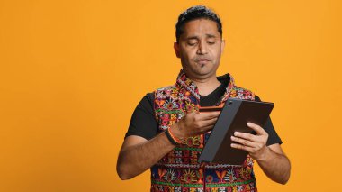 Man shakes head in disbelief after realizing lack of necessary funds while trying to shop online on tablet. Disappointed indian person gets credit card transaction rejected, studio backdrop, camera A clipart