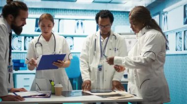 Kabinede test sonuçları ve sağlık kayıtlarını karşılaştıran uzman doktorlar, hastalık önleme konusunda meslektaşlarına danışıyorlar. Muhtelif sağlık görevlileri bir toplantıda profesyonel uzmanlıklarını paylaşıyorlar. Kamera B.