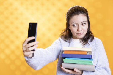 Gülümseyen kadın okul sınavı için bilgi içeren kitap yığınıyla telefon özçekimi yapıyor. Stüdyo geçmişi. Öğrenci üniversite değerlendirmesinden önce akıllı telefonuyla fotoğraf çekiyor, ders kitaplarını tutuyor