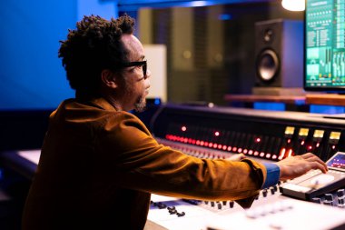 Afrikalı Amerikalı müzik yapımcısı kontrol masasındaki ses ayarlarını ayarlıyor, profesyonel stüdyodaki melodileri düzenliyor. Müzik mühendisi müzik düzenlemek için konsolu karıştırma, faders itme üzerinde çalışıyor.