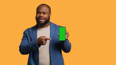 Gülümseyen Afro-Amerikan adam cep telefonunu yeşil ekran ile sunuyor, stüdyo arka planında izole edilmiş. Neşeli BIPOC çalışanı, boş fotokopi odası, kamera A ile promosyon yaratıyor.