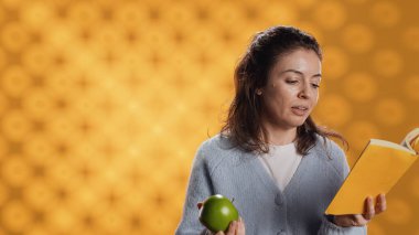 Kitap okuyan, kol kaslarını çalıştırmak için havaya elma atan, sağlıklı yaşayan, stüdyo arka planında izole edilmiş bir kadın. Roman ve yeşil meyvelerle eğlenen, aktif olan kişi
