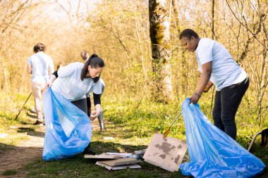 Ormanı çöpten temizlemeye çalışan çeşitli gönüllüler, doğal ekosistemi korumak için kirlilikle mücadele ediyorlar. Genç aktivist takımı aletlerle çöp ve plastik atık topluyor..