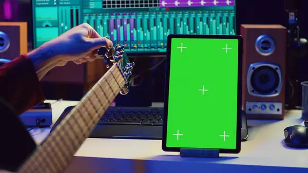 stock image Audio engineer practices tuning his acoustic guitar in home studio, watching a video tutorial online via tablet with greenscreen. Musician learning to play instrument and create music. Camera A.