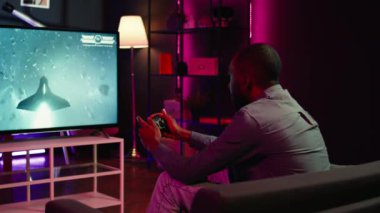 Oyuncu, akıllı televizyonlarda birinci şahıs nişancı video oyunu oynamak için kulaklık ve kumanda kullanıyor. Ev sinemasındaki adam düşmanları yenmek için yüksek teknoloji oyun konsolunu kullanıyor, kamera B