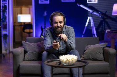 Keyifli bir oyuncu evde oyun oynuyor ve keyfine bakıyor. Neon ışıklı oturma odasında kanepede oturan neşeli adam, konsolda video oyunu oynuyor, yoğun bir iş gününden sonra dinleniyor.