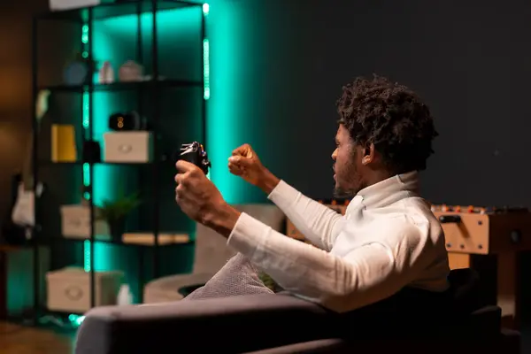 Stock image African american gamer throwing fists in air from joy after finally finishing difficult videogame. Man feeling excited after defeating enemies during gaming session on console in neon lit living room