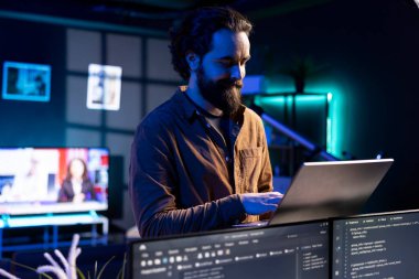Gülümseyen teknisyen dizüstü bilgisayar kullanarak yazılım hata ayıklaması yapıyor, PC monitörlerinde gösterilen kod satırlarındaki arızaları kontrol ediyor. Mutlu adam evine girip defter kullanarak kodlama sorunlarını hallediyor.