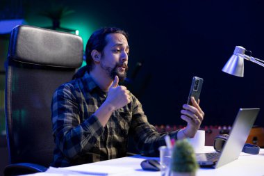 Serbest çalışan bir erkek müşterisiyle telefonda video konferans görüşmesi yapıyor. Sakallı adam dizüstü bilgisayarının önünde oturuyor, ailesi ve arkadaşlarıyla sanal bir toplantı için mobil cihazını kullanıyor..