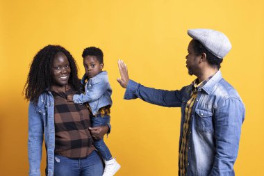 Afrikalı Amerikalı baba küçük çocuğuna ve karısına el sallıyor, sarı arka planda buluşuyorlar. Genç siyahi aile stüdyoda poz veriyor, baba küçük çocukla eğleniyor..