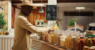 Afrikalı Amerikalı müşteriler makarna ya da kavanozlarda tahıl alışverişi yapıyor, yeniden kullanılabilir konteynırlarda depolanan çeşitli ürünlere bakıyor. Çevre dostu işleri destekleyen, kirletici olmayan ürünler alan biri. Kamera 2.