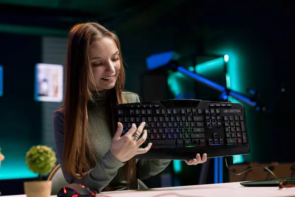 stock image Joyous girl reviews RGB lit keyboard from sponsor for online streaming channel in apartment. Smiling media star excited to promote gaming computer peripherals brand in neon lit studio