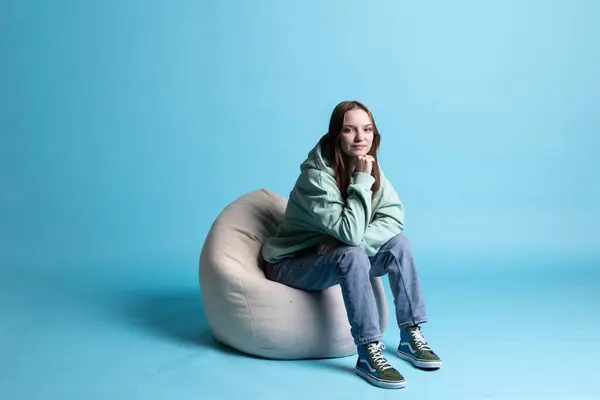 stock image Portrait of happy girl sitting on beanbag armchair and smiling, isolated over blue studio background. Upbeat teenager seated on cozy bean bag, resting at home, having positive mood, studio backdrop