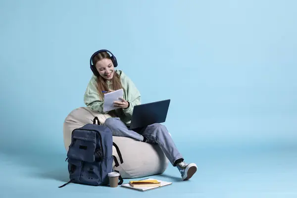 stock image Smiling girl using laptop to learn information for high school exams while listening music, studio background. Upbeat teenage student hearing music and holding notebook, writing on notepad