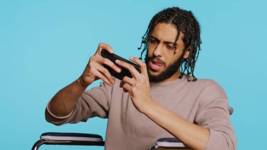 Özürlü bir adam cep telefonuyla yoğun video oyunu oynar, dokunmatik ekranda spam saldırı tuşları kullanır. Oyuncu tekerlekli sandalyede, stüdyo geçmişinde, B kamerasında oyun yarışmasına katılıyor.