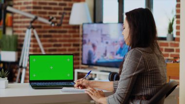 Evde kalan tele-işçi, ahşap masadaki çeşitli işleri yapmak için yeşil ekran laptopunu kullanıyor. Krom tuşlu klavyeye veri yazıp not defterine yazan uzak çalışan bir çalışan