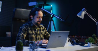 Gülümseyen adam ofis masasında oturuyor ve dizüstü bilgisayarda klavye çalıyor, görevleri çözüyor. Oturma odasından iş ortaklarına e-posta göndermek için dizüstü bilgisayar kullanan mutlu bir işçi, kamera B