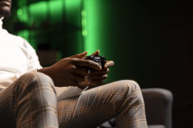 Man holding controller in green neon illuminated apartment, playing videogames. Gamer on couch using joystick to participate in PvP online multiplayer game on console system, relaxing and having fun clipart