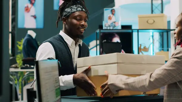 stock image Retail employee picking up clothes order boxes from department store, signing invoice and confirmation papers at cash register counter. African american woman giving requested merchandise. Camera A.