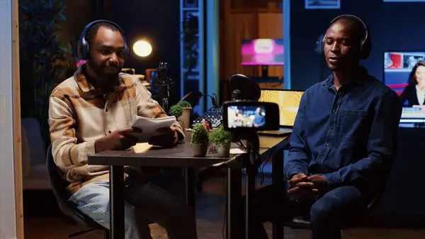 stock image Content creator recording interview with professional camera in studio, reviewing notes to ask pertinent questions to guest. Journalist using filming gear, talking on mic with african american man