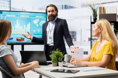Finansal danışman büyük ekranda müşterilere istatistiksel grafikler gösteriyor, piyasa eğilimlerini analiz ederek geliri optimize ediyor. Yatırım planını kontrol eden aile üyeleri danışman tarafından sergileniyor