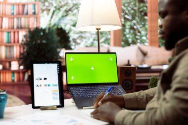 Küçük işletme sahibi dizüstü bilgisayarında fotokopi alanı ile mali raporlar üzerinde çalışıyor, bilgi grafikleriyle muhasebe analizi yaptıktan sonra notlar alıyor. Yönetici evde yeşil ekran kullanıyor.