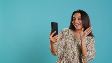 Mutlu kadın, arkadaşlarıyla sohbet etmek için internetten akıllı telefondan arkadaşça jestler yapıyor. Telekonferansta gülümseyen Hintli insan cep telefonu, stüdyo arka planı, kamera A 'yı kullanıyor.