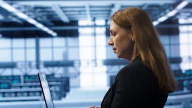 Veri merkezindeki sistem yöneticisi dizüstü bilgisayarda yazıyor, sorunları düzeltmek için hata kontrol araçları çalıştırıyor. Sunucu merkezindeki kadın, araç gereçlerini etkileyen ağ sorunları arıyor. Not defteri cihazı, kamera A 'yı kullanarak.