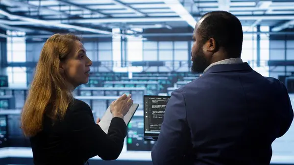 stock image Teamworking programmers in data center overseeing server operations using laptop and tablet. Coworkers ensuring optimal performance and data integrity in server room using devices, camera A