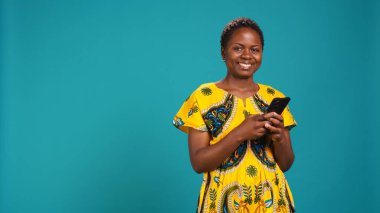 Native girl in ethnic outfit browsing internet webpages in studio, using her smartphone to check texts and social media platforms. Young person standing against blue background. Camera B. clipart