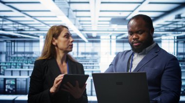 Teamworking technicians managing data center with advanced technology using devices. Coworkers in server room optimizing data center infrastructure with laptop and tablet, camera B clipart