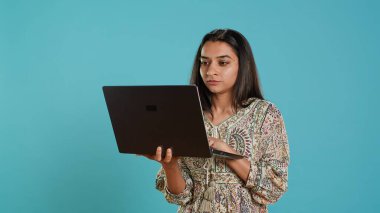 Indian woman typing on laptop keyboard and cursing, feeling angry at work. Person writing emails on notebook, complaining loudly, isolated over studio background, camera A clipart