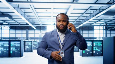 Bilgi işlem teknisyeni veri merkezinde telefonla konuşuyor, sunucu bakımını ve sorun çıkarma görevlerini koordine ediyor. Sunucu rafları ve yüksek teknolojili ekipmanlarla çevrili bir yönetici. Telefon görüşmesi, kamera B