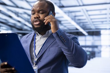 Afrikalı Amerikalı bir adam veri merkezi sistemlerinin kesin belgelerini tutuyor, çalışanlarla telefonda konuşuyor. Denetmen çağrı sırasında sunucu odası çalışma alanı ayarları ve prosedürleri hakkında konuşur