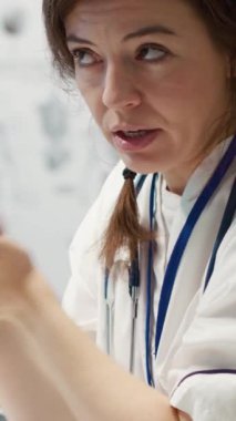 Dikey Görüntü Hekimi, bir kabinede tıbbi sonuçları tartışmak için bir hastayla buluşur. Röntgen taramalarını yorumlamak için ileri seviye teşhis cihazları kullanır ve profesyonel bir sağlık yaklaşımı gösterir.