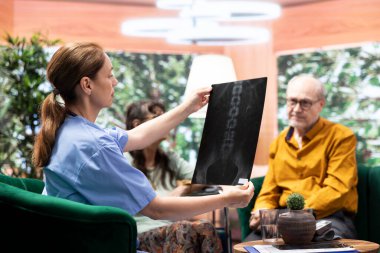 Hemşire, yaşlı hastalara teşhisi anlamak için radyografi sonuçlarını açıklıyor. Yaşlı kadın tekerlekli sandalye kullanıcısı iyileşmek için mücadele ediyor. Tıbbi asistan ziyaret sırasında destek öneriyor.