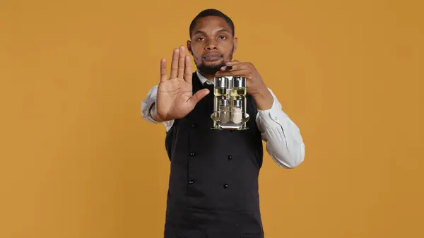 stock image Waiter in uniform showing a stop sign with his palm raised in studio, expressing negative symbol and refusal about something. Showing rejection and being dissatisfied. Camera B.