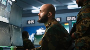 Asker askeri üste görevler yapıyor, dijital radar ekipmanlarıyla gözetleme yapıyor, füze sistemleri çalıştırıyor. Ortadoğulu bir adam, ordu merkezinin güvenlik kameralarına izinsiz hareketlilik var mı diye bakıyor. Kamera A.