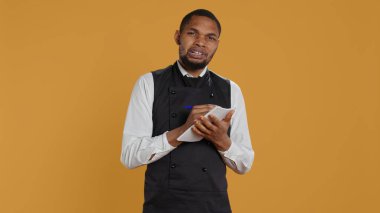 Profesyonel yemek şirketi müşterilerine fatura için not defteri hazırlıyor. Sarı arka planda istenen yiyecekleri not alıyor. Önlüklü garson, müşteri memnuniyeti için defter servisi. Kamera B.