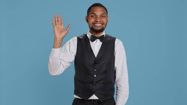 Otel müşterilerine gülümseyen ve el sallayan belboy çalışanı, insanları özgüvenle selamlıyor ve keyifli bir konaklama için lüks hizmetler sunuyor. Neşeli personel insanlara merhaba diyor. Kamera B.