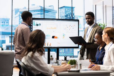 Kurul odasındaki konferans masasında işbirliği yapan iş ortakları, pazar eğilimleri ve ekonomik tahminlerin ayrıntılı bir analizini sunmak için akıllı teknoloji ve aylık istatistiklerden yararlanıyorlar..