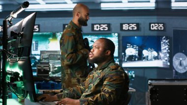 Askeri ajan kontrol odasında çalışıyor, hava keşif verilerini analiz ediyor ve meslektaşımla konuşuyor. İstihbarat elemanı ekranları kontrol ediyor, iş arkadaşı, kamera A ile tartışıyor.