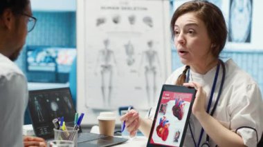 Kalp fonksiyonlarını ve durumunu test sonuçlarına göre açıklayan bir kadın uzmanı, kabinedeki doktor hasta etkileşimi. Doktor kardiyolog kardiyovasküler sistemi tablette gösteriyor. Kamera A.