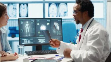Afro-Amerikalı doktor bir hastaya röntgen taraması sonuçlarını sunuyor. Tıbbi kabinedeki radyografi taramalarını inceliyor. Doktor sağlık için ilaç öneriyor. Kamera A.