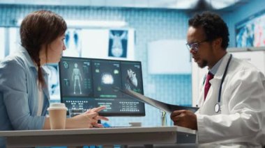 MRI sonuçlarına ve röntgen görüntülerine dayanarak doktordan gelen teşhis raporunu dinleyen kadın hasta tıbbi dolaptaki bilgisayara işaret ediyor. Afrikalı Amerikalı uzman yardım ediyor. Kamera A.