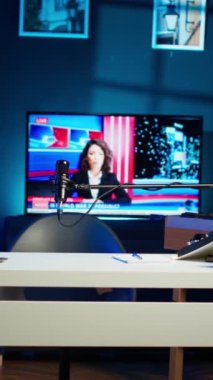 Ev içi boş dizüstü bilgisayar ve ev bitkileriyle dolu dikey video çalışma masası. Oturma odası neon ışıklarıyla aydınlatılıyor. Masada bir defter var. Televizyonda haber yayını var.