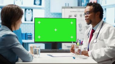 Afro-Amerikan sıhhiyeciler, hastasına danışıp sağlık sigortası ile ilgili tedavi planlarını incelerken bilgisayardaki yeşil ekrana işaret ediyorlar. Doktor hasta kadına tıbbi tavsiye veriyor. Kamera A.